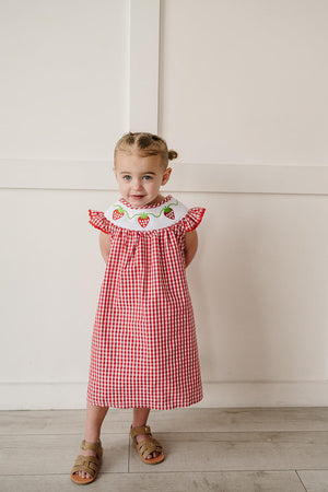 A young girl stands smiling in a red and white gingham dress with a ruffled collar adorned with embroidered strawberries.