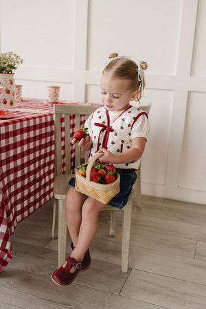 A young girl sits on a wooden chair, holding strawberries while wearing a white blouse with red polka dots and a red bow.