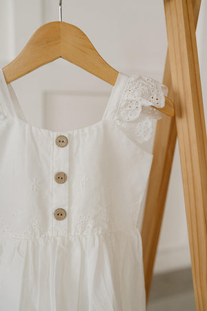 A close-up of a white cotton dress featuring delicate eyelet detailing and wooden buttons, hanging on a wooden hanger against a light background, showcasing its elegant design for children.
