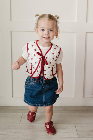 A toddler wearing a strawberry-printed quilted vest smiles while walking on a light wooden floor. The outfit includes a denim skirt and red shoes, set against a neutral wall.