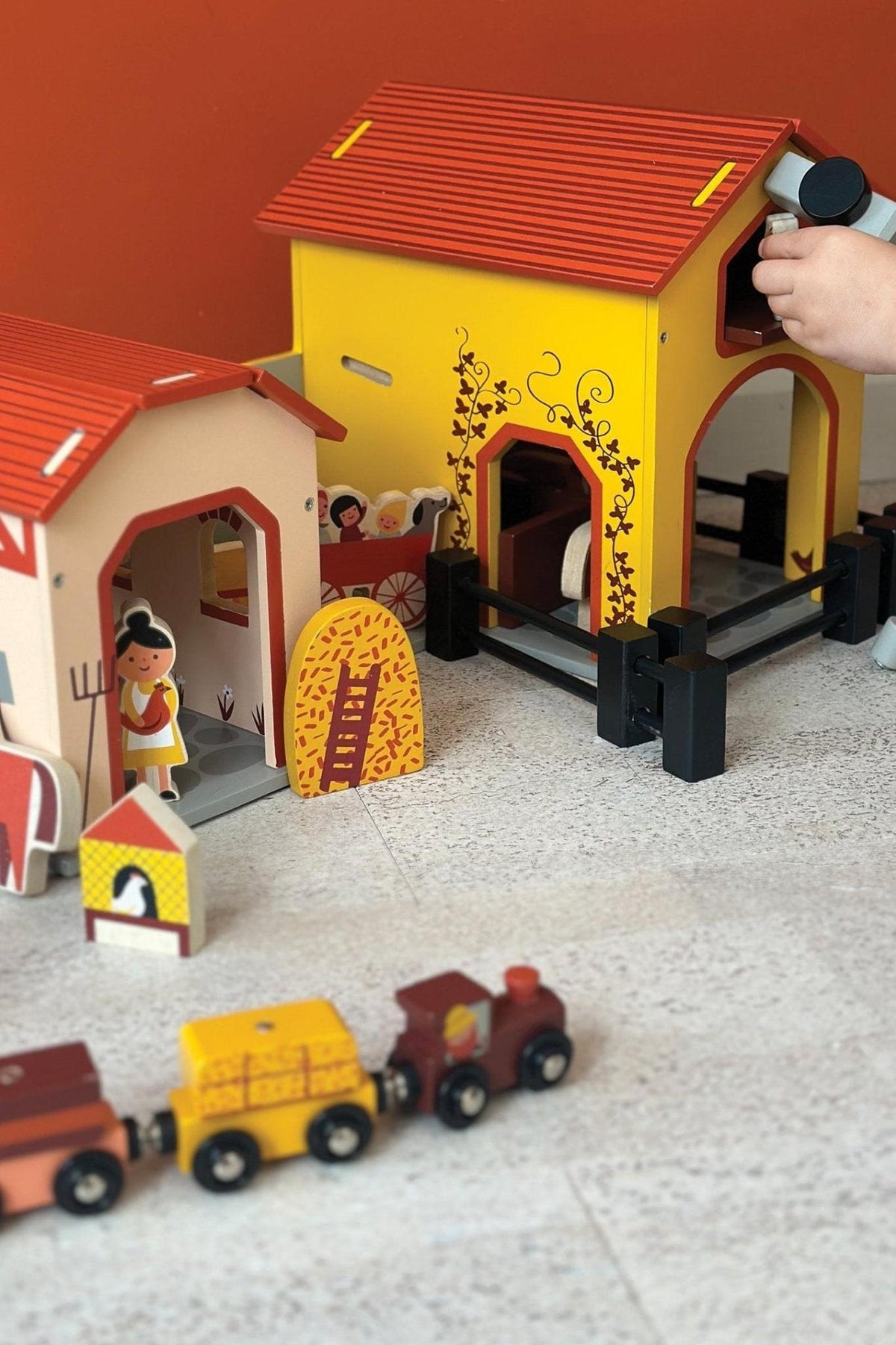 Colorful wooden farm set with barns, fences, and a toy train on a light floor. Childs hand visible.