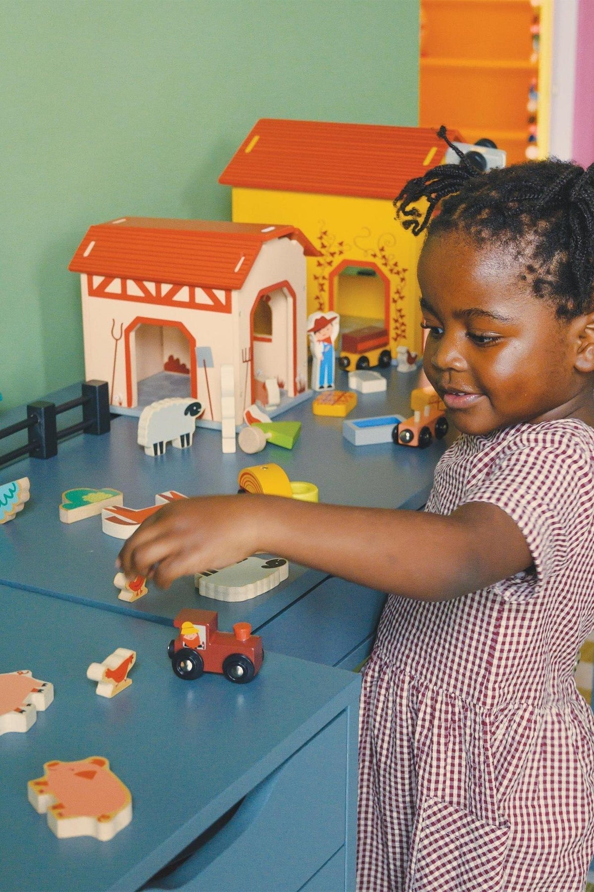 A child plays with colorful wooden farm toys on a blue table, featuring barns, animals, and a tractor.