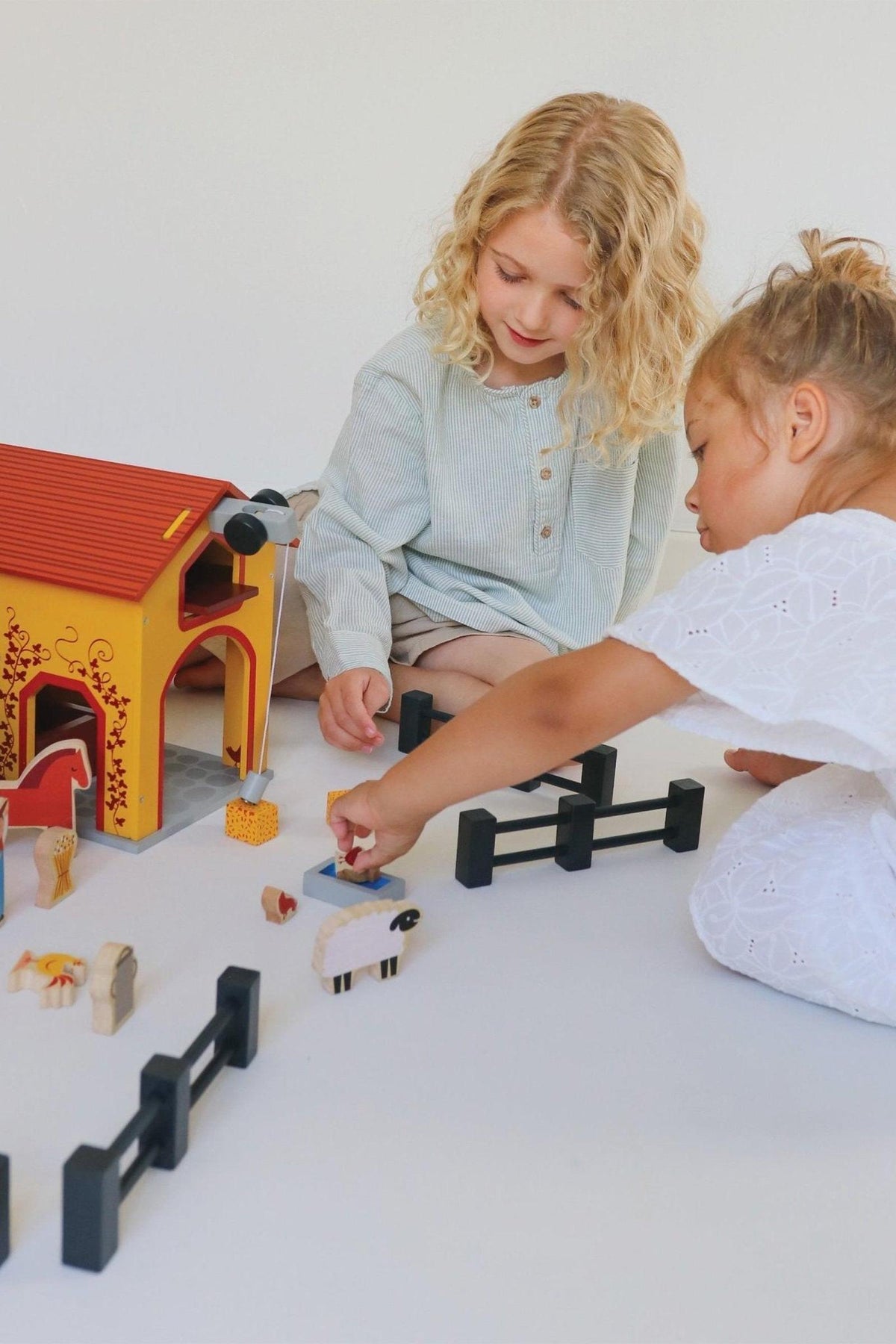 Children play with a brightly colored wooden barn set, featuring animals and fences on a white surface.