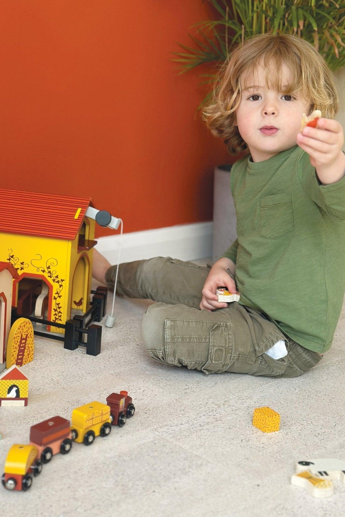A child in green plays with a colorful wooden toy barn and train set on a light floor.