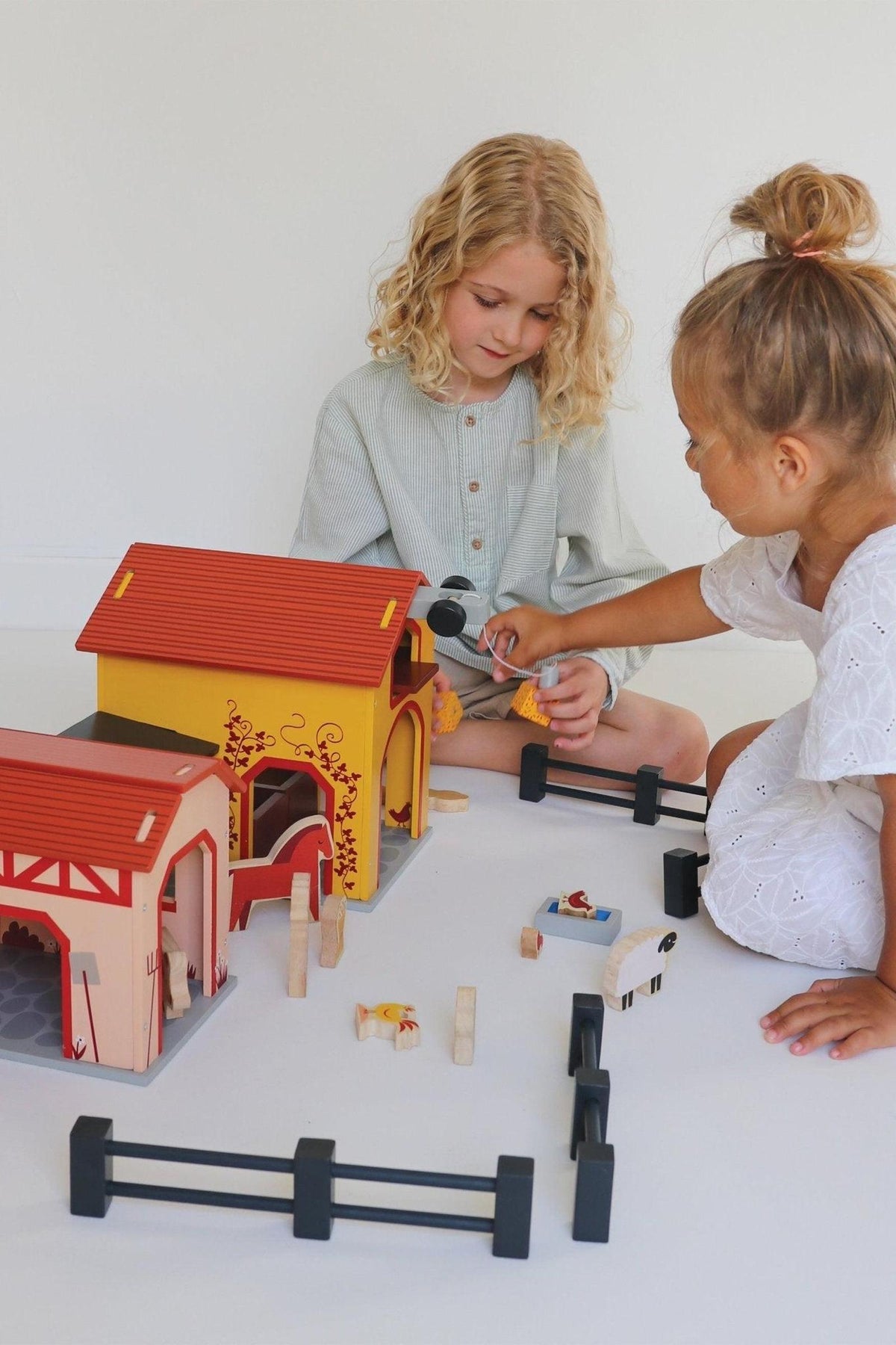 Two children play with a colorful wooden farm set featuring barns, animals, and fences on a white surface.