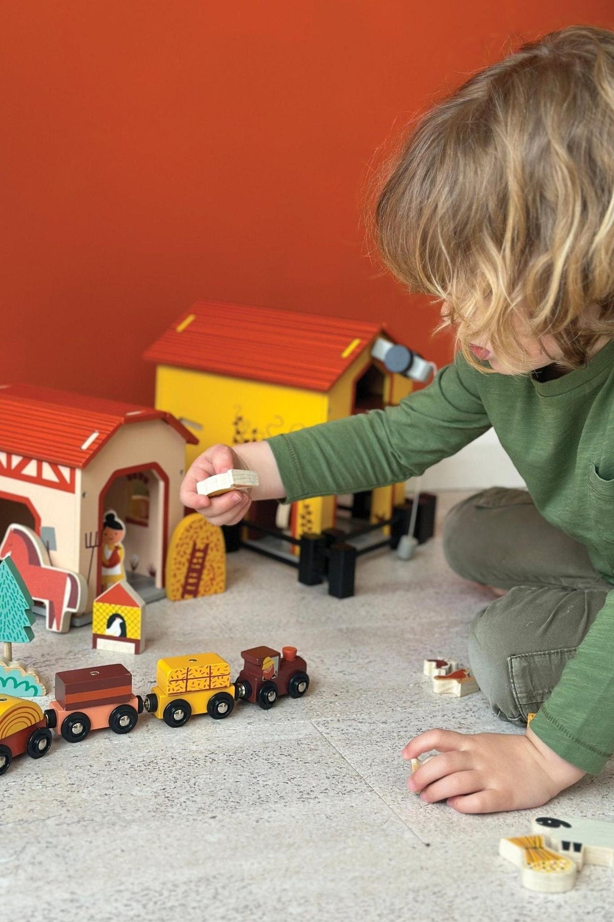 A child plays with a colorful wooden farm set, featuring barns and toy vehicles on a light surface.