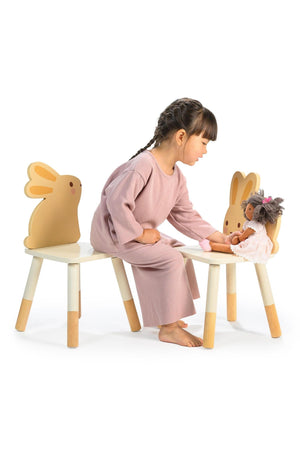 A child in a pink outfit plays with a doll on a bunny-themed chair set with wooden legs.