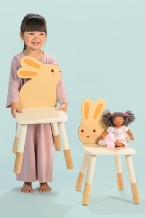 A child holds a bunny-eared chair from a toddler furniture set, with another chair featuring a doll beside it.