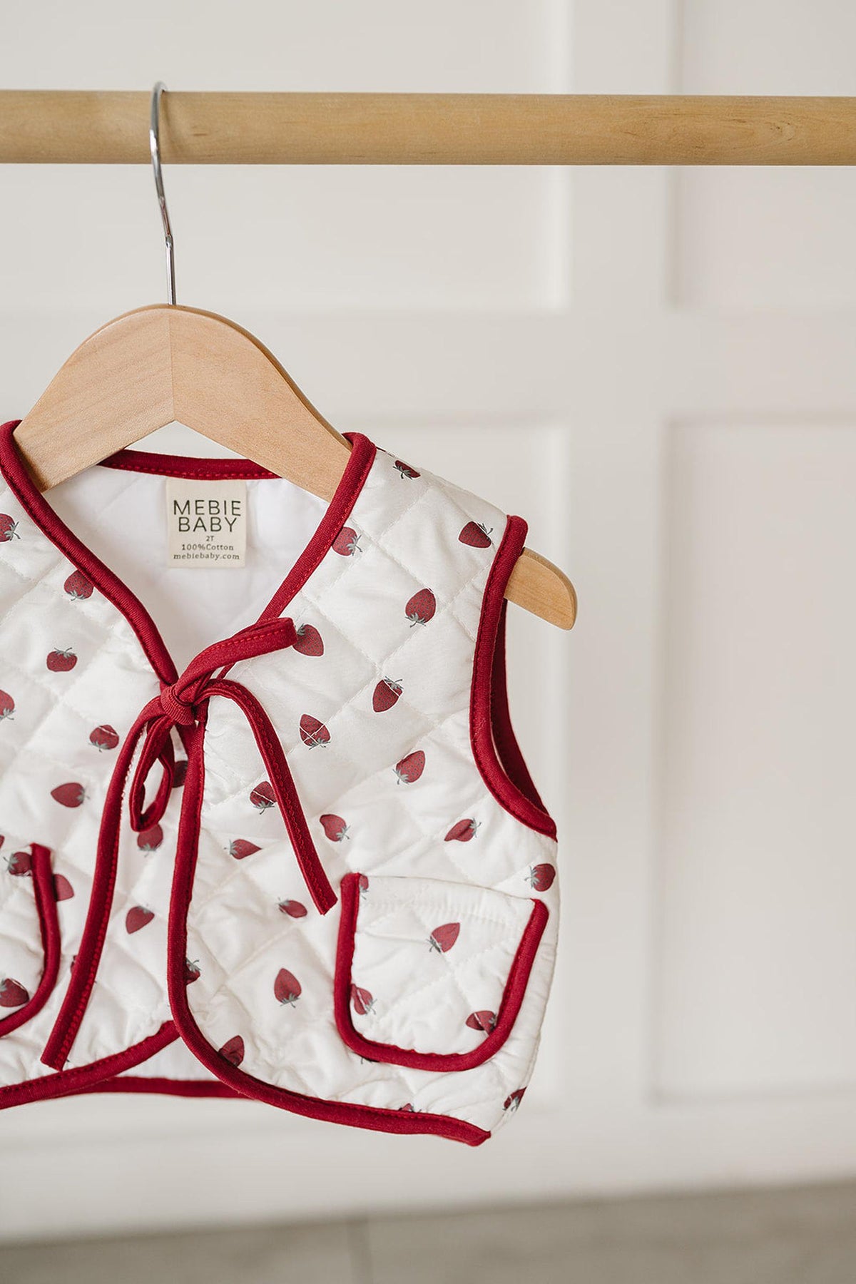 A quilted cotton vest from Mebie Baby, featuring a cheerful allover strawberry print and a red trim. The vest is displayed hanging on a wooden hanger against a neutral background.