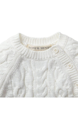 Close-up of a soft white cable knit sweater romper by Hope & Henry, featuring button openings at the shoulder. Made from 100% organic cotton, its designed for babies comfort.