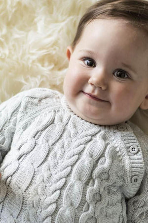 A smiling baby lies on a soft surface, wearing a light gray cable knit sweater romper made from organic cotton.