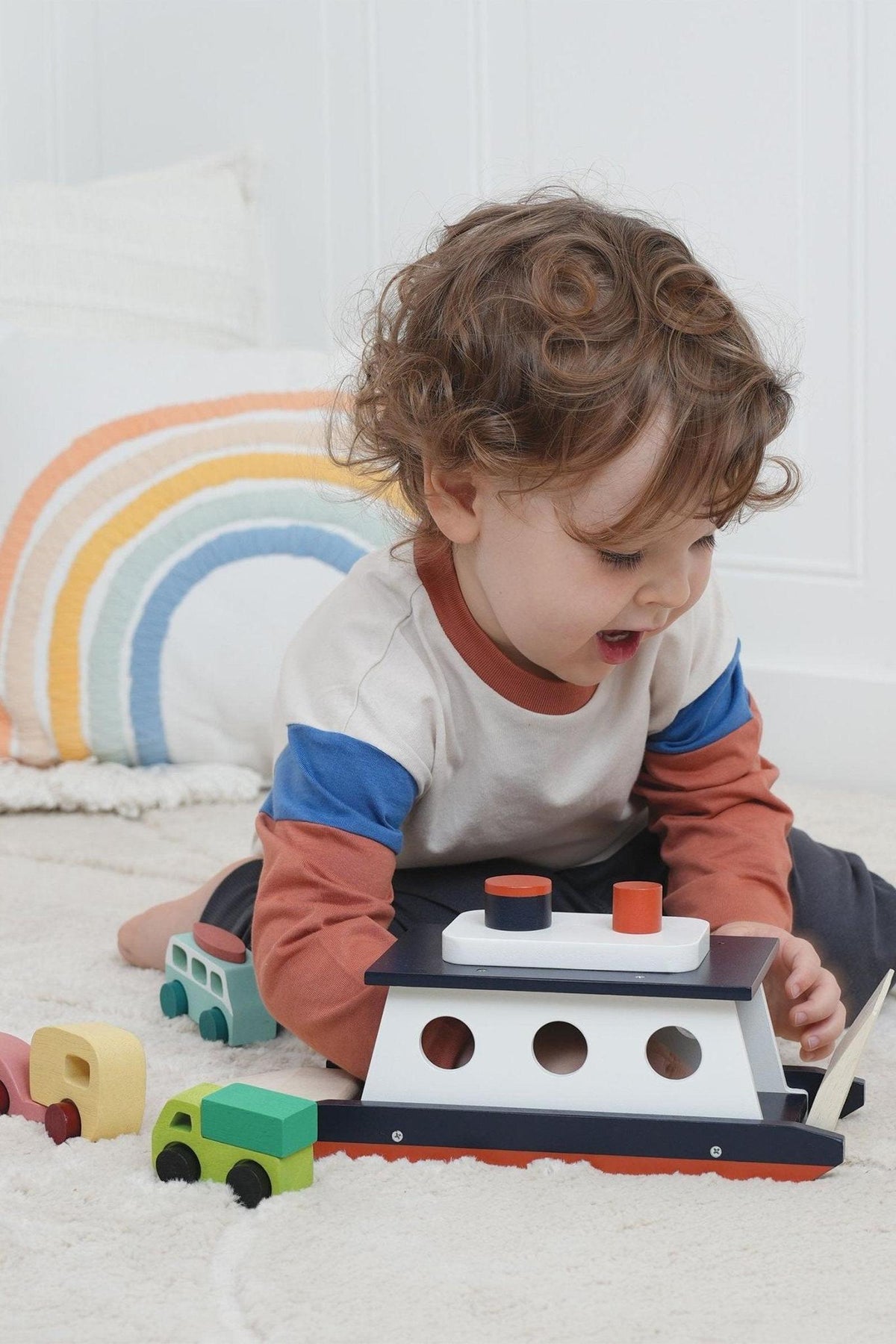 A child plays with a wooden toy ferry and colorful cars on a soft, white carpet with a rainbow pillow.