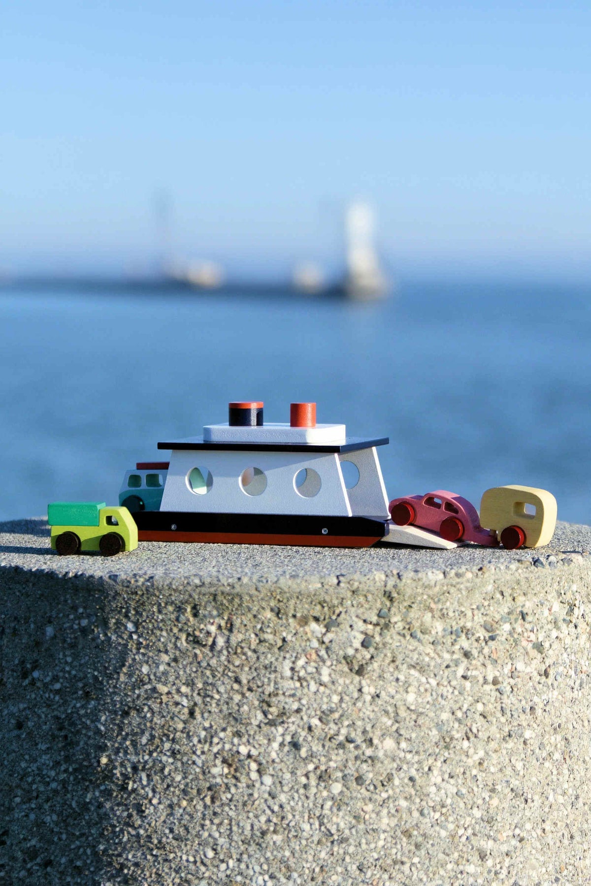 Wooden toy ferry with cars on a concrete surface, set against a blurred ocean background under a clear sky.