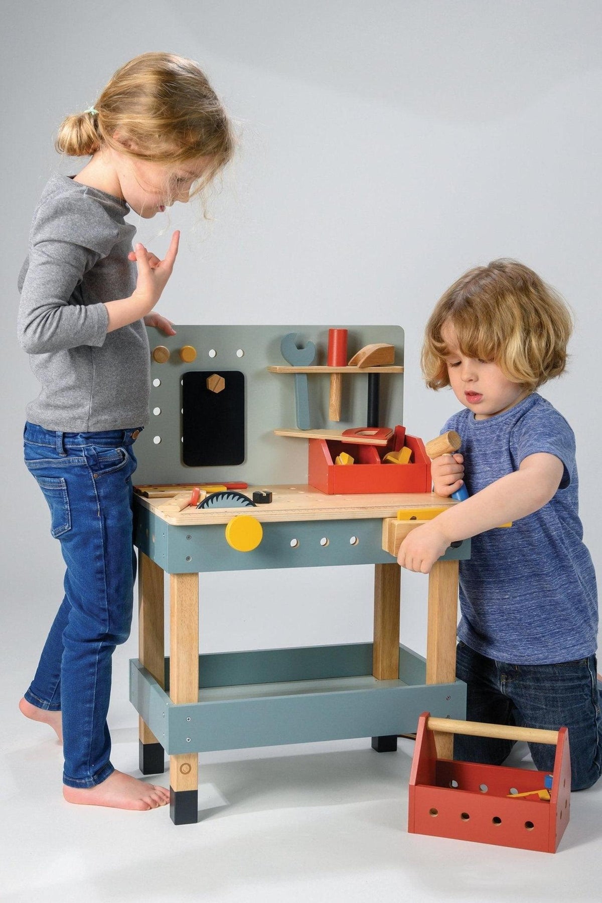 Two children playing with a wooden carpenters workshop, featuring tools and a red toolbox, on a light background.