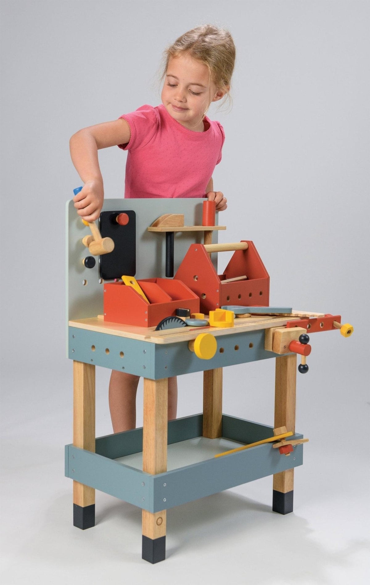 A child in a pink shirt plays with a colorful wooden carpenters workshop toy, featuring tools and accessories.
