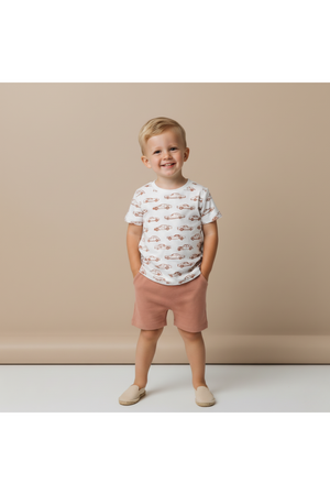 A smiling child stands against a neutral backdrop, wearing a white tee featuring colorful car prints and soft, pink shorts.