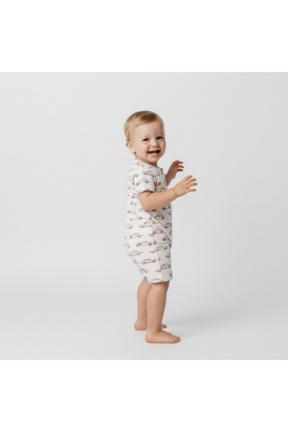 A joyful toddler wearing an organic cotton short zip romper with a playful car pattern stands against a plain white background.