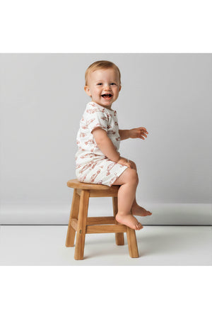 A cheerful toddler sits on a wooden stool, wearing a white organic cotton romper featuring a playful car print. The soft fabric is suitable for young children and promotes comfort.