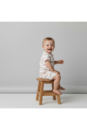 A happy toddler sits on a wooden stool, wearing an organic cotton short zip romper with a playful car pattern. The soft, neutral background highlights the rompers charming design.