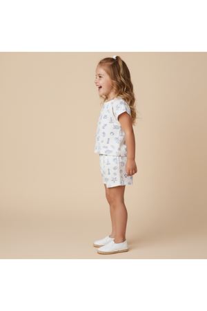 A young girl stands smiling in a coastal-themed organic cotton drop shoulder tee and shorts set, featuring playful nautical prints. The soft, lightweight fabric is ideal for warm days.
