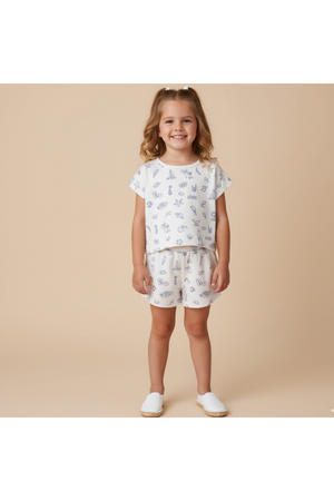 A young girl stands smiling in a light-colored, organic cotton drop shoulder tee and shorts set, featuring playful seaside-themed prints.