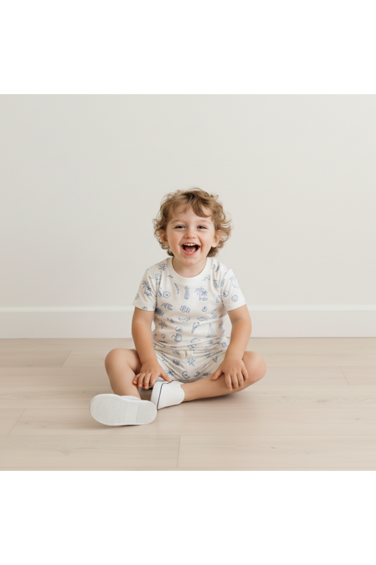 A smiling child sits on a light wooden floor, wearing a white organic cotton tee and shorts set featuring a playful coastal print. The relaxed fit emphasizes comfort and fun.