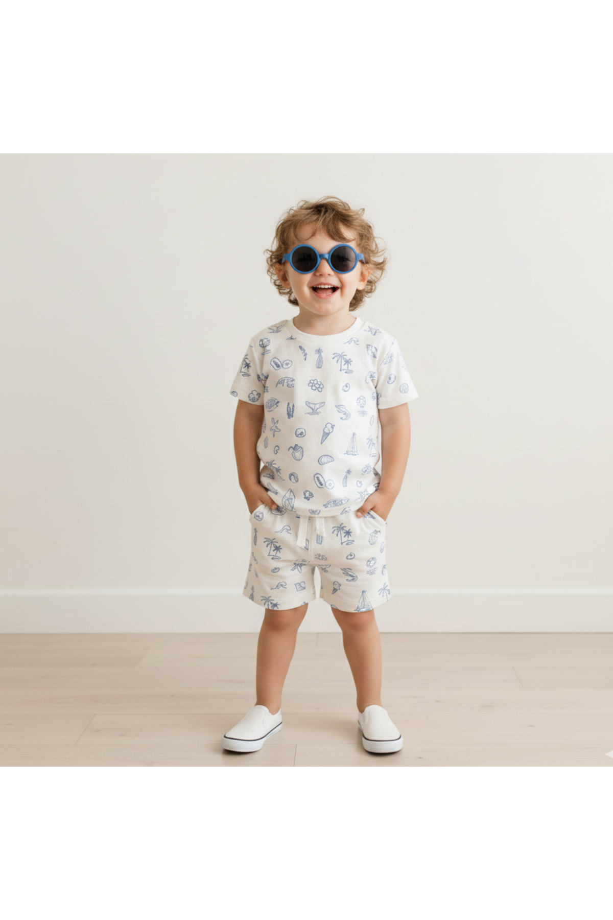 A smiling child wearing a Coastal print organic cotton tee and shorts set stands against a light wall. The outfit features playful beach-themed designs, perfect for summer activities.