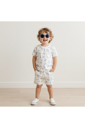 A smiling child wearing a Coastal print organic cotton tee and shorts set stands against a light wall. The outfit features playful beach-themed designs, perfect for summer activities.