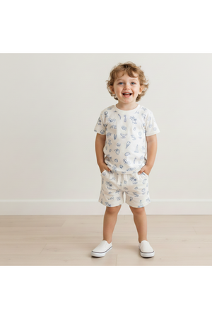 A cheerful young child stands smiling in a light-colored, organic cotton tee and shorts set featuring playful coastal prints. The outfit is perfect for warm weather and casual play.