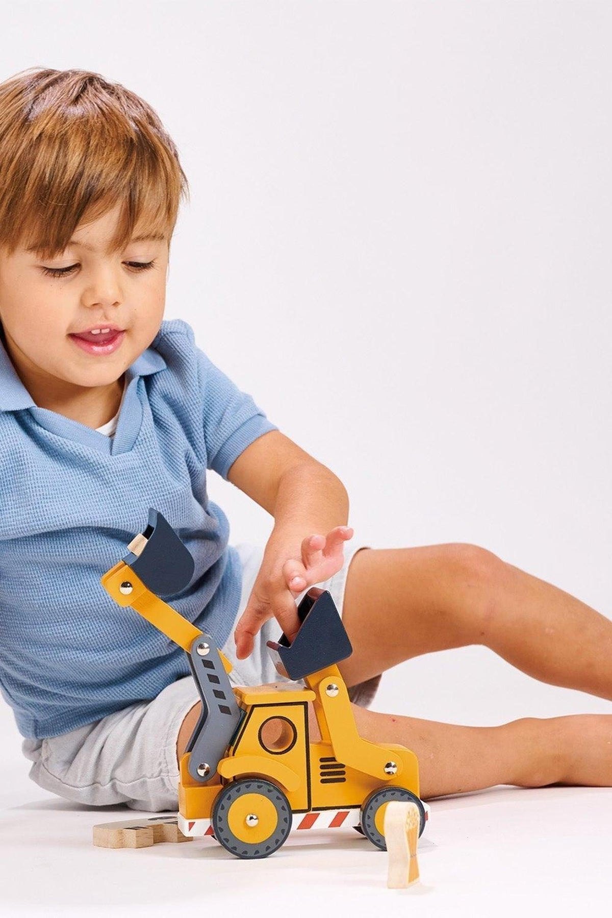 A child in a blue shirt plays with a yellow wooden backhoe toy on a white surface.