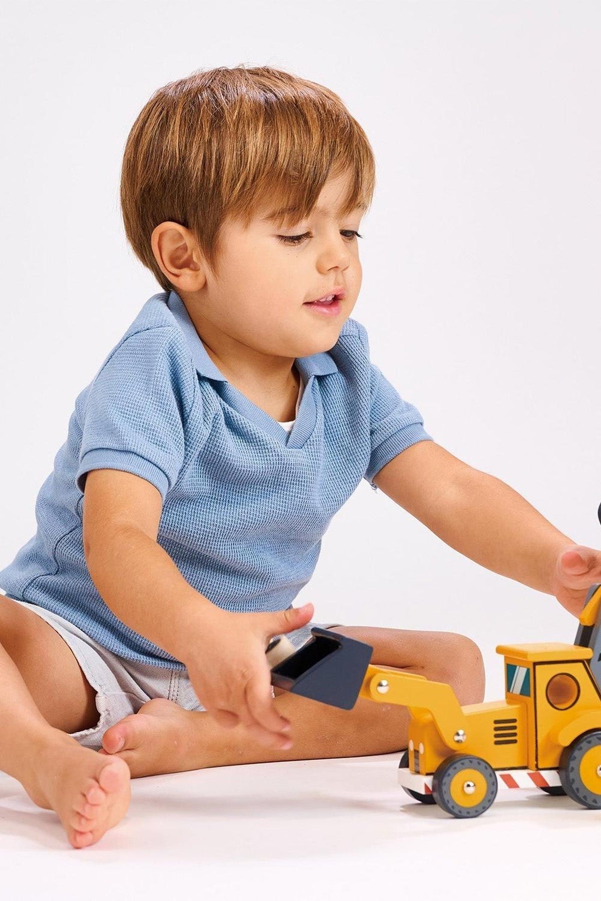 A young child in a blue shirt plays with a yellow wooden toy backhoe on a white background.