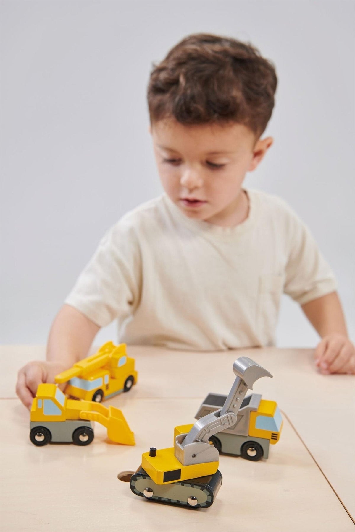 A child plays with yellow and gray wooden construction vehicles, including a backhoe and excavator, on a table.