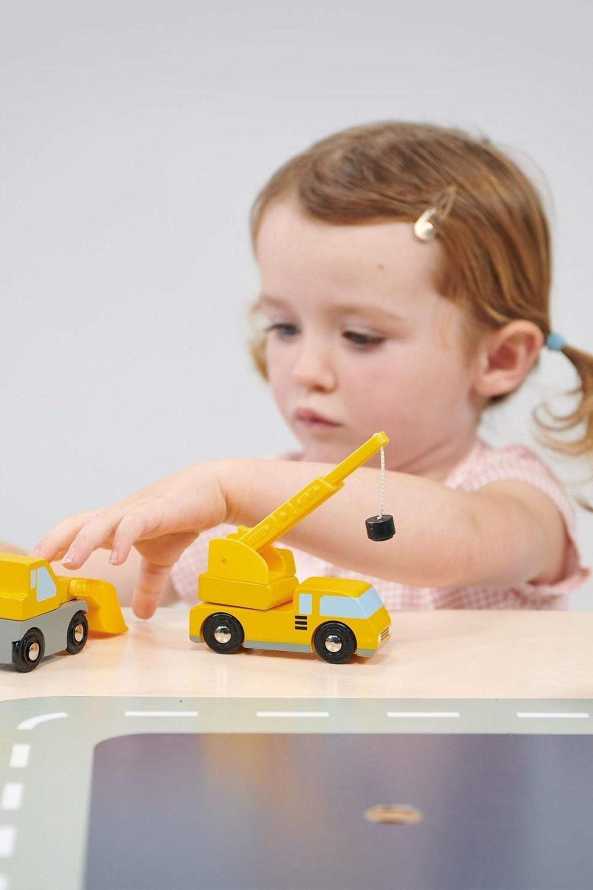 A child plays with yellow wooden toy construction vehicles on a table, focusing on a crane and a truck.