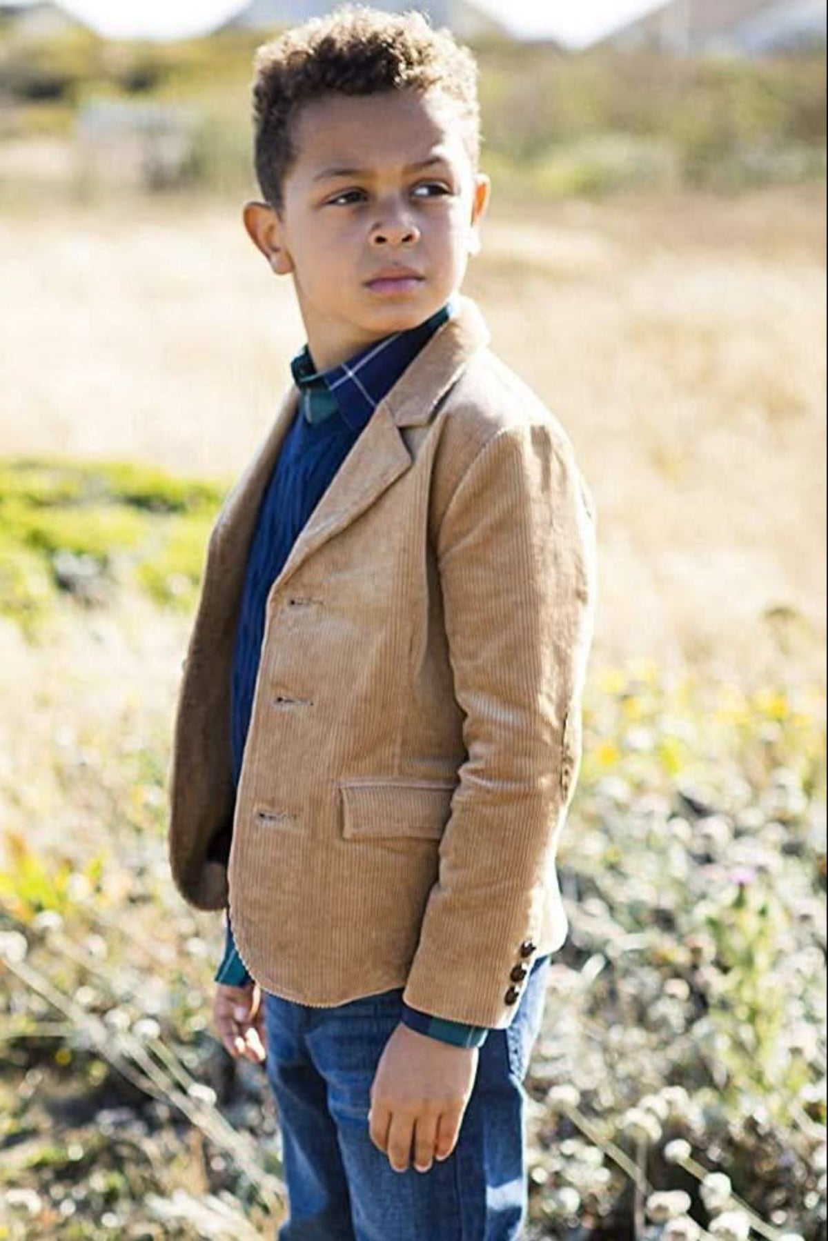 A young boy stands in a field wearing a medium brown corduroy blazer over a blue shirt and plaid collar.