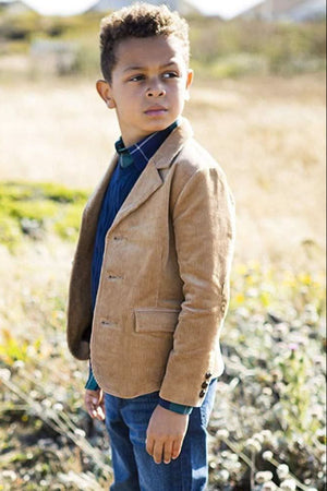 A young boy stands in a field wearing a medium brown corduroy blazer over a blue shirt and plaid collar.