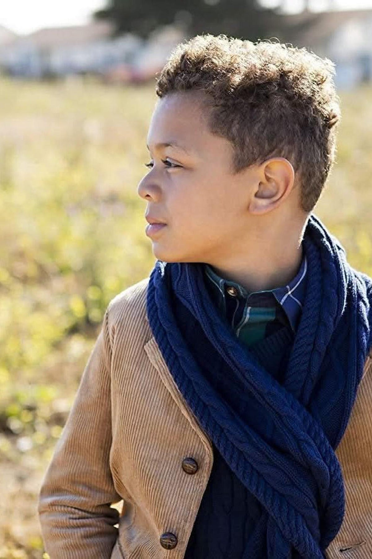 A young boy stands outdoors, wearing a medium brown corduroy blazer and a navy scarf. His curly hair and thoughtful expression complement the natural, sunny setting in the background.