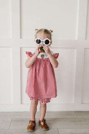 A young child wears a red gingham dress with ruffled sleeves and a strawberry embroidered neckline, paired with matching bloomers and oversized round sunglasses, standing against a white paneled wall.