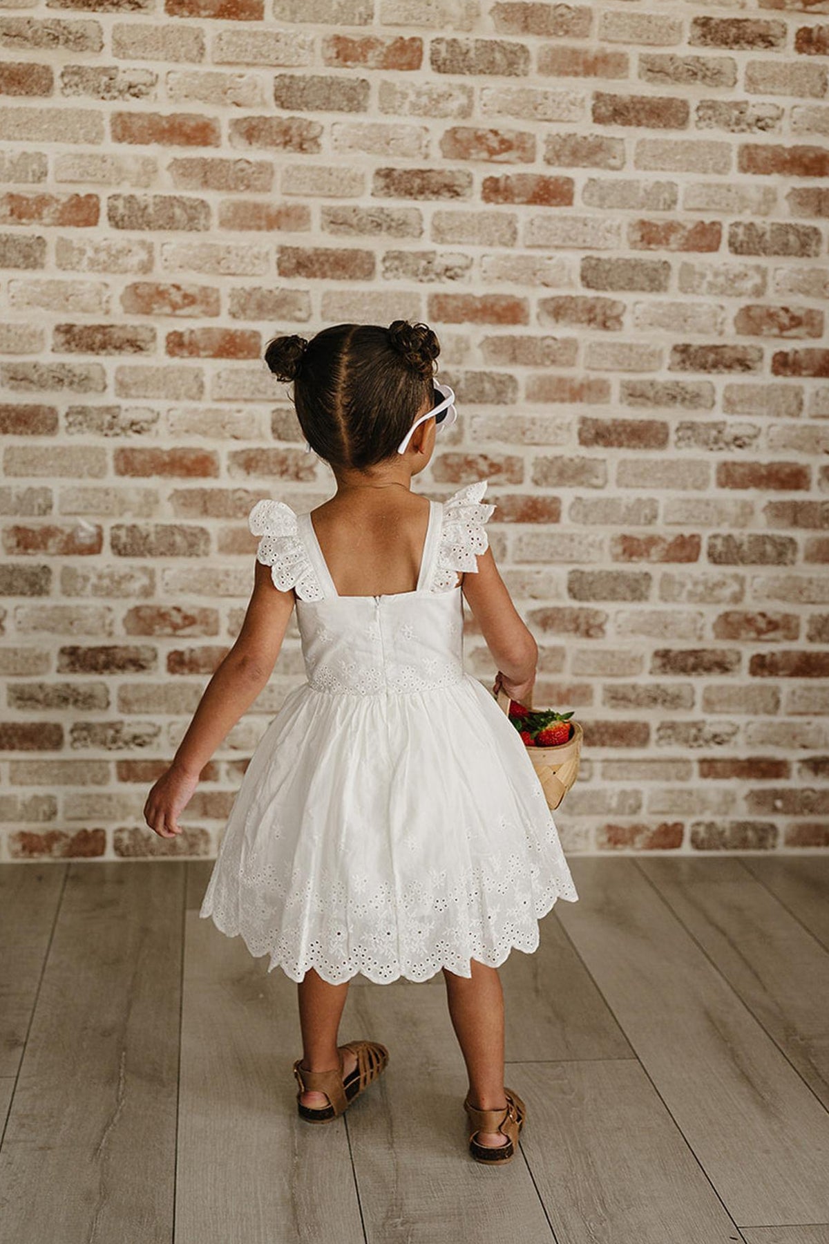 A young child stands with their back to the camera, wearing a white eyelet cotton dress with ruffled sleeves.