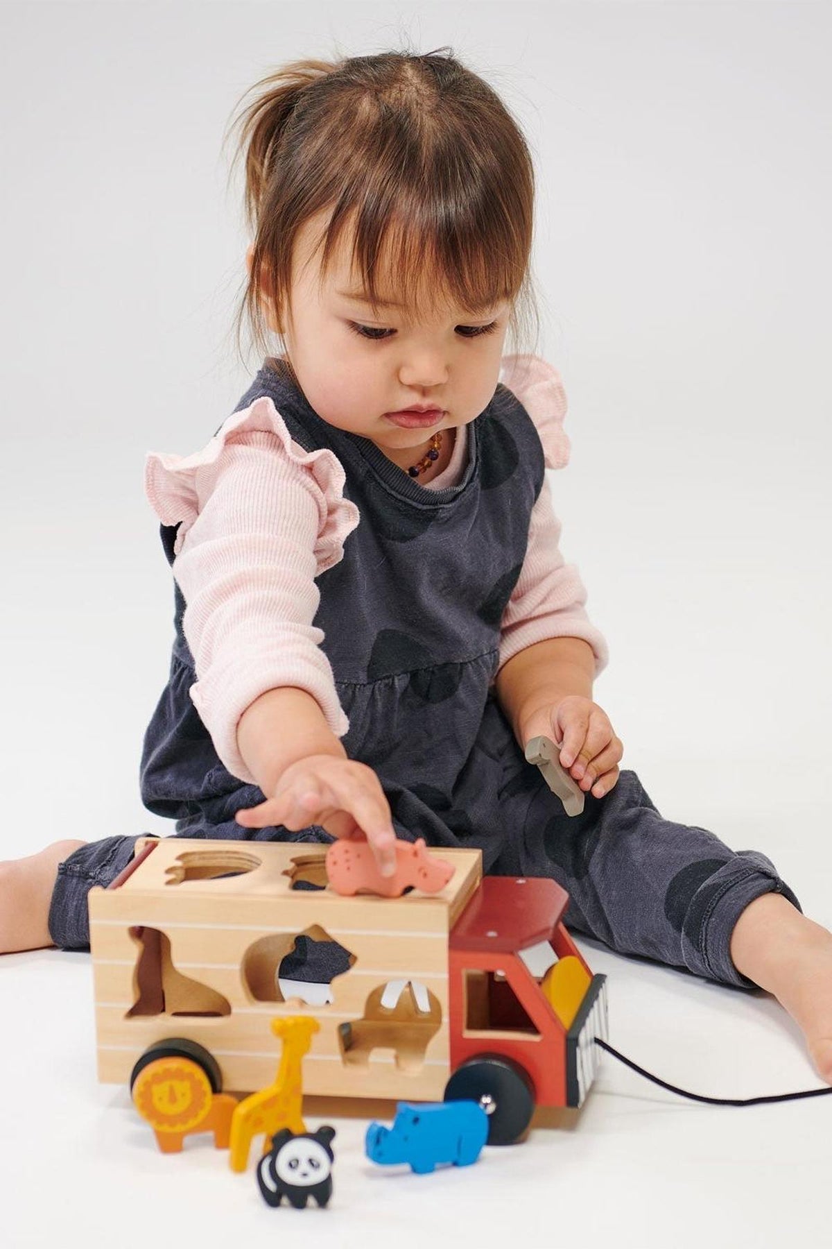 A toddler plays with a wooden safari truck shape sorter, featuring colorful animal pieces on a white background.