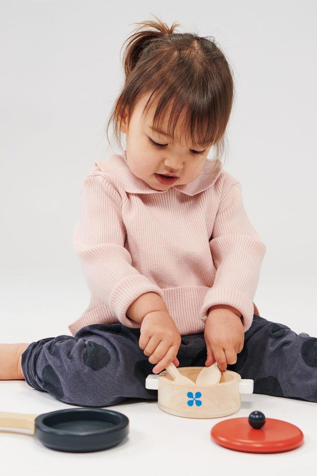 A toddler in a pink sweater plays with a wooden toy pan set, featuring a pot and lid.