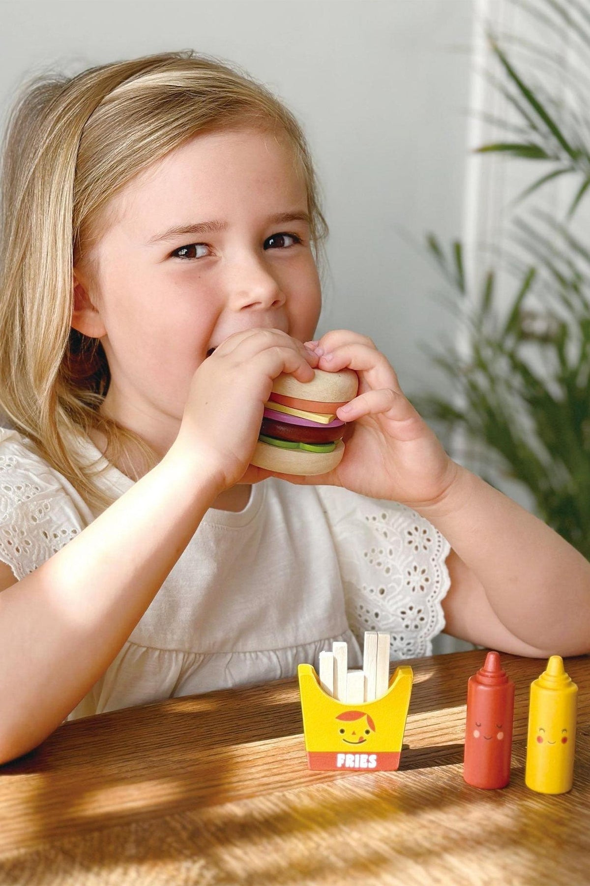 A child in a white shirt plays with a wooden burger toy, fries, and condiments on a wooden table.