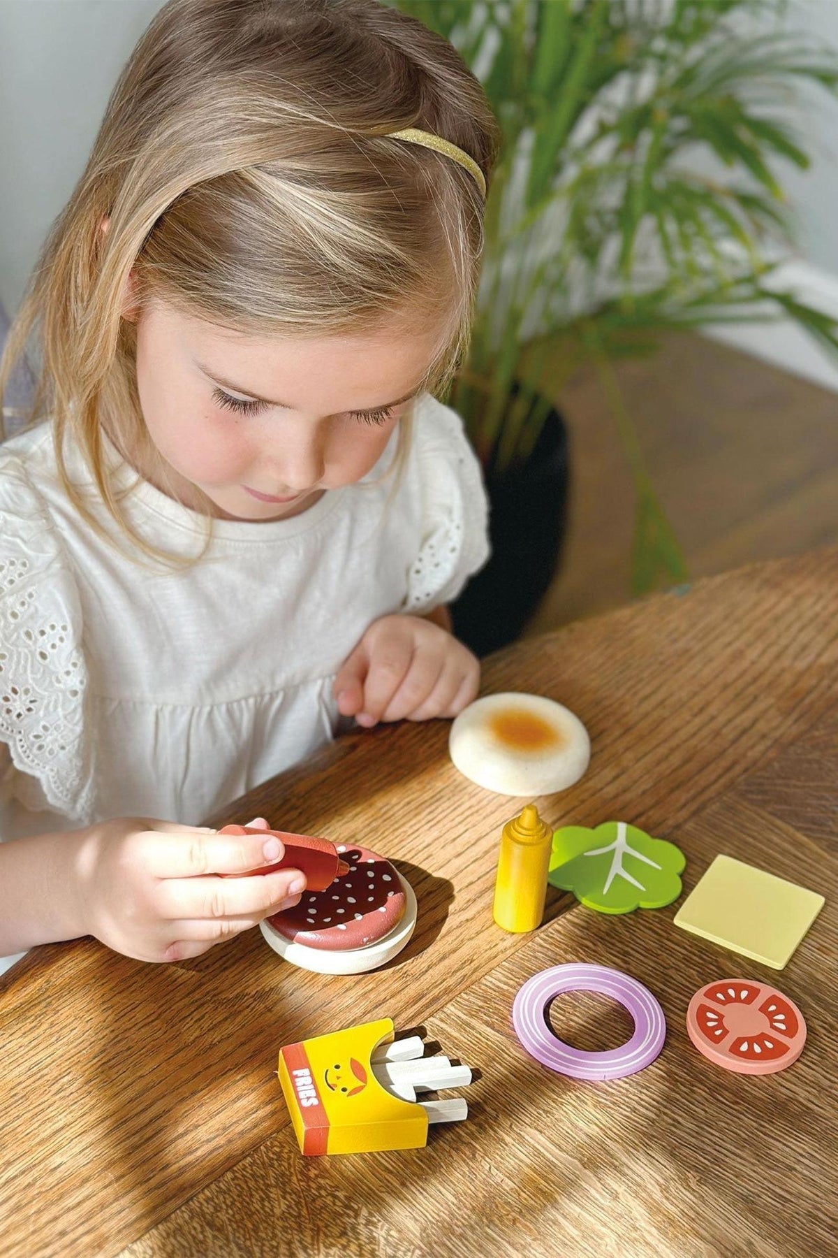 A child plays with a wooden fast food toy set, including a burger, fries, and toppings, on a table.
