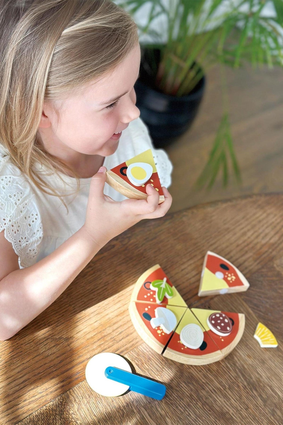 A child plays with a wooden pizza toy set on a wooden table, featuring colorful toppings and a pizza cutter.