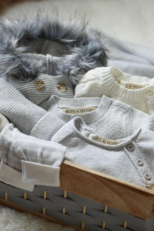 A cozy assortment of Hope & Henry baby sweaters in light gray and cream is neatly arranged in a wooden.