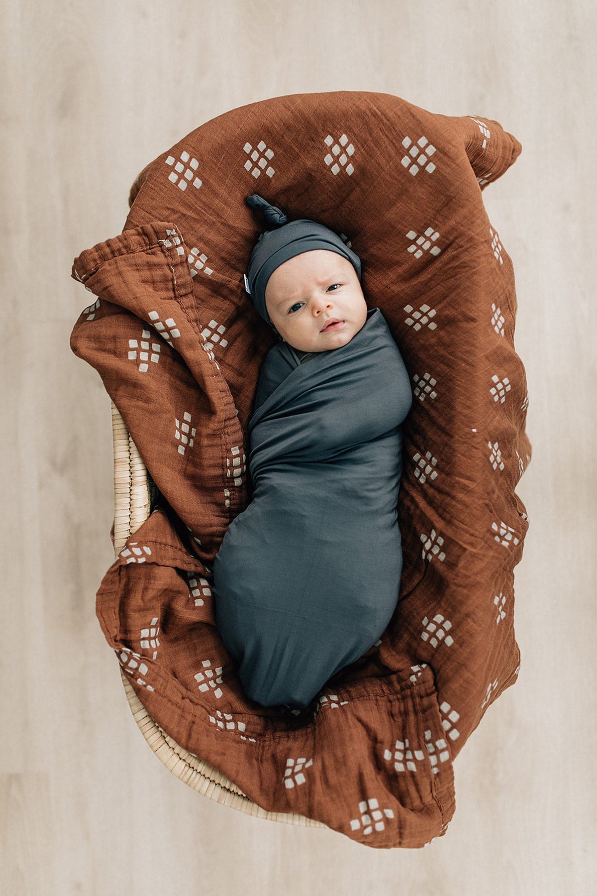 A newborn in a charcoal knot hat and swaddle lies on a patterned brown blanket in a basket.