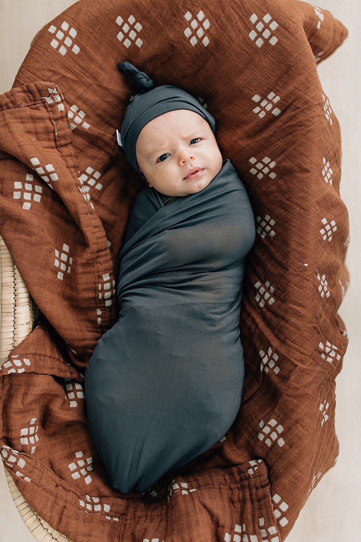 A baby wrapped in a charcoal swaddle and matching hat, lying on a brown patterned blanket.