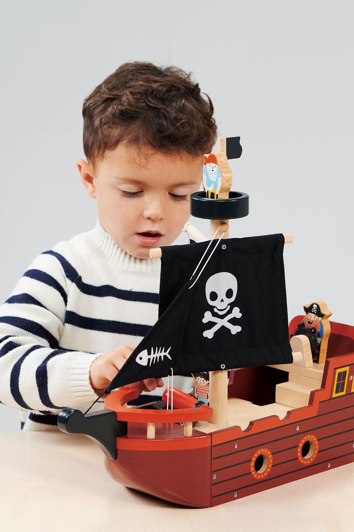 A child plays with a wooden pirate ship featuring a black skull-and-crossbones sail and colorful pirate figures.