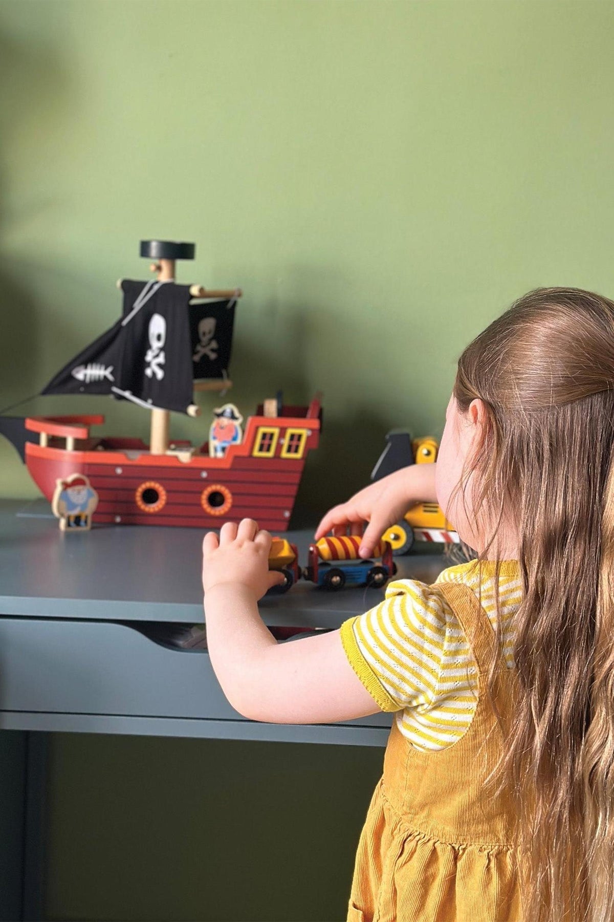 A child plays with a colorful wooden pirate ship and toy cars on a gray table.