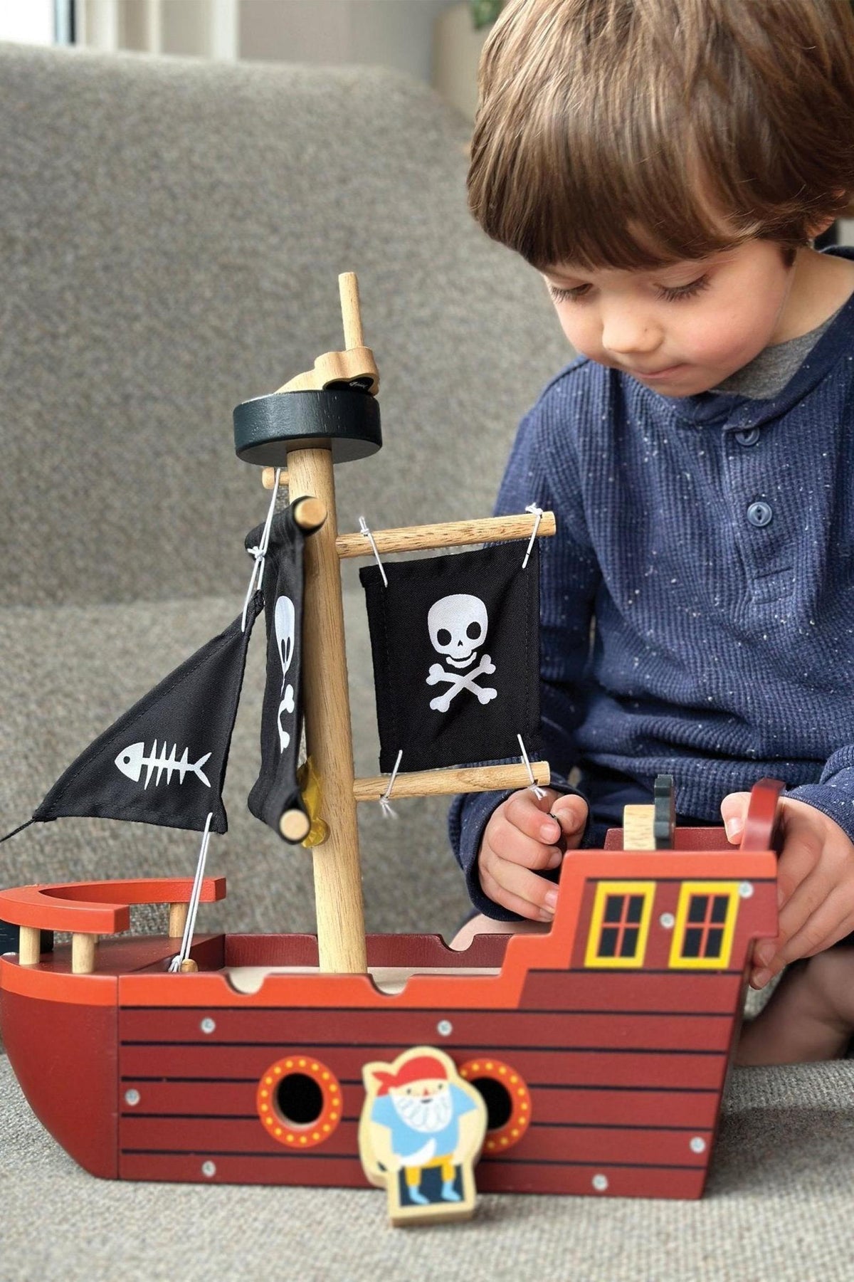 A child plays with a wooden pirate ship featuring black sails with skull and fishbone designs, indoors.
