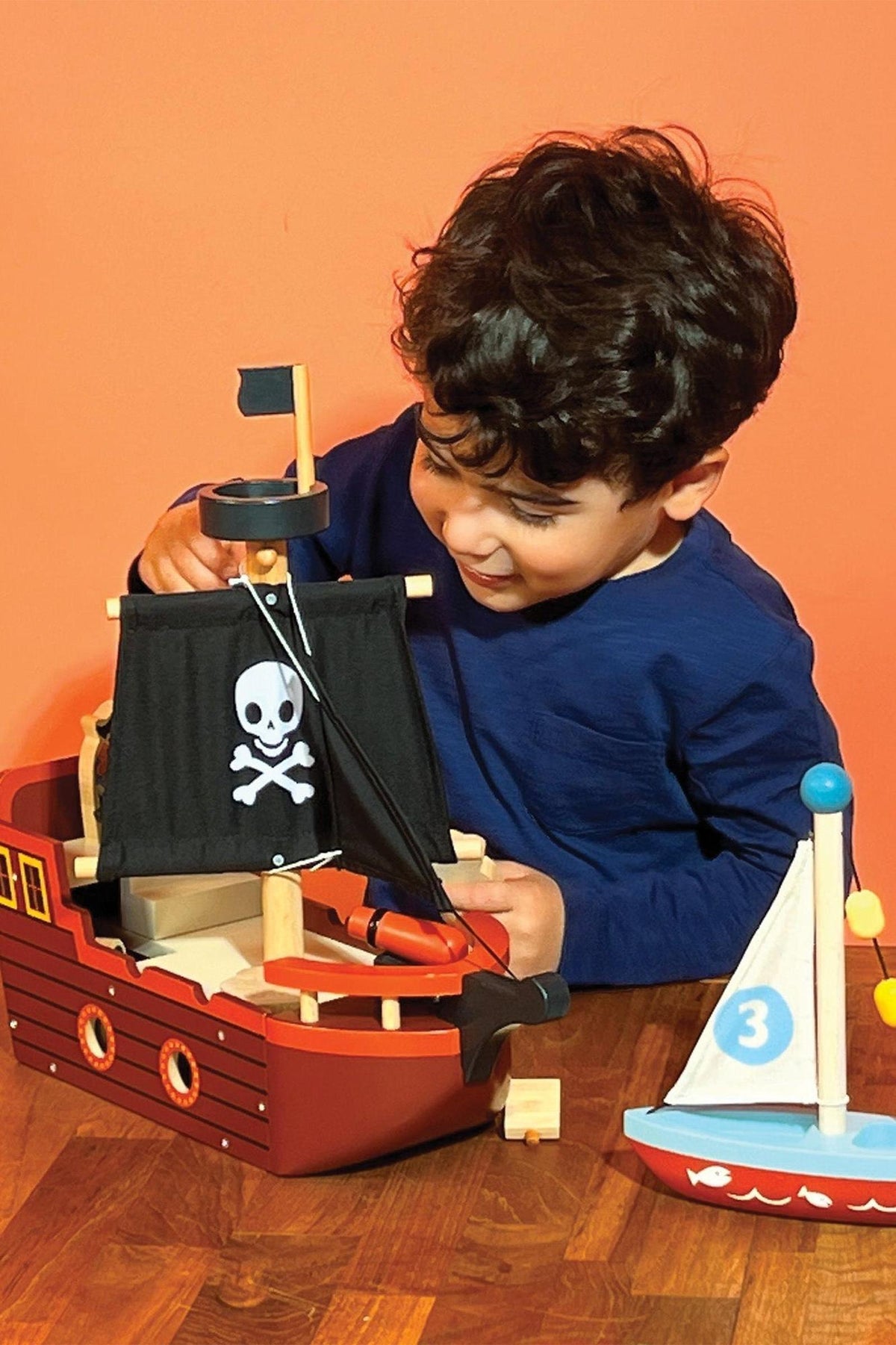 A child plays with a wooden pirate ship featuring a black sail with a skull and crossbones design.